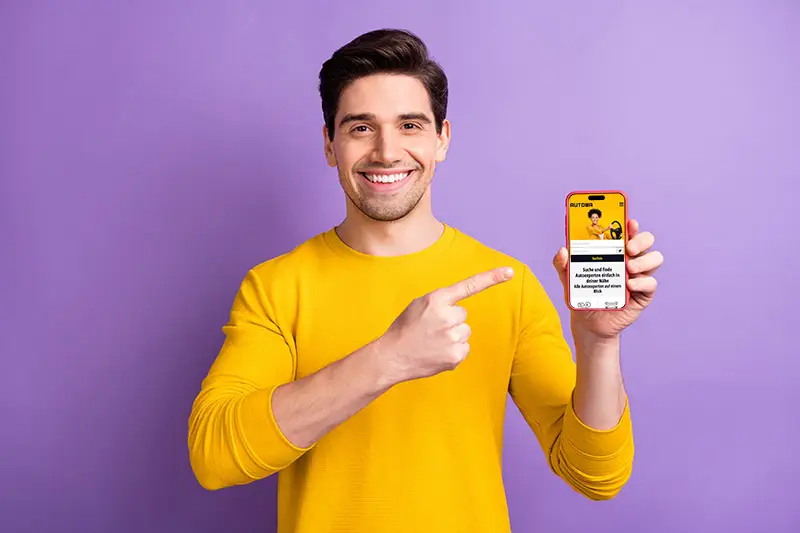 Autoba - Ein Mann im gelben Oberteil hält einen Smartphone in der linken Hand und zeigt mit dem rechten Zeigefinger auf dem Smartphone.